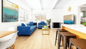 A modern living room with a blue sofa, a coffee table, and a TV. There is a kitchen area with a white countertop and a sink. - NEW! Ocean View Beach House Steps from the Sand  (Manhattan Beach)