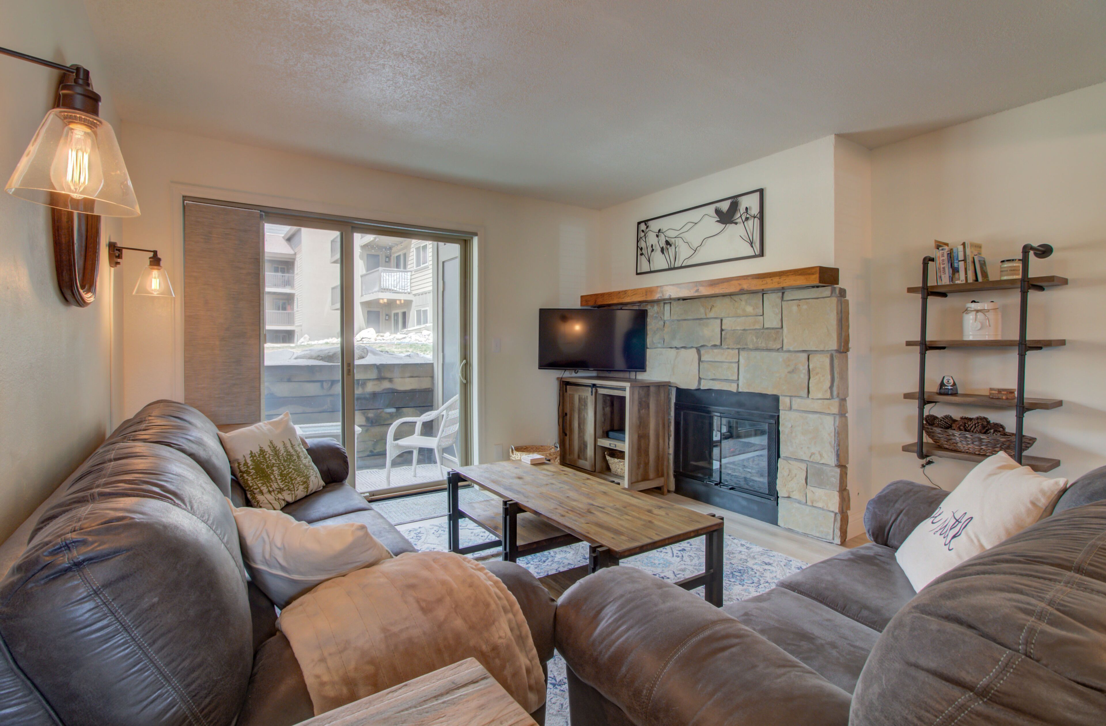 A cozy living room with leather sofas, a stone fireplace, a flat-screen TV, and a large sliding glass door leading to a balcony.