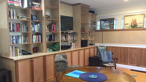 A living room with a wooden bookshelf filled with books and DVDs, a round wooden coffee table with a placemat, and a chair with a patterned cushion. - Country Blue is close to forest trails and beach (Qualicum Beach)