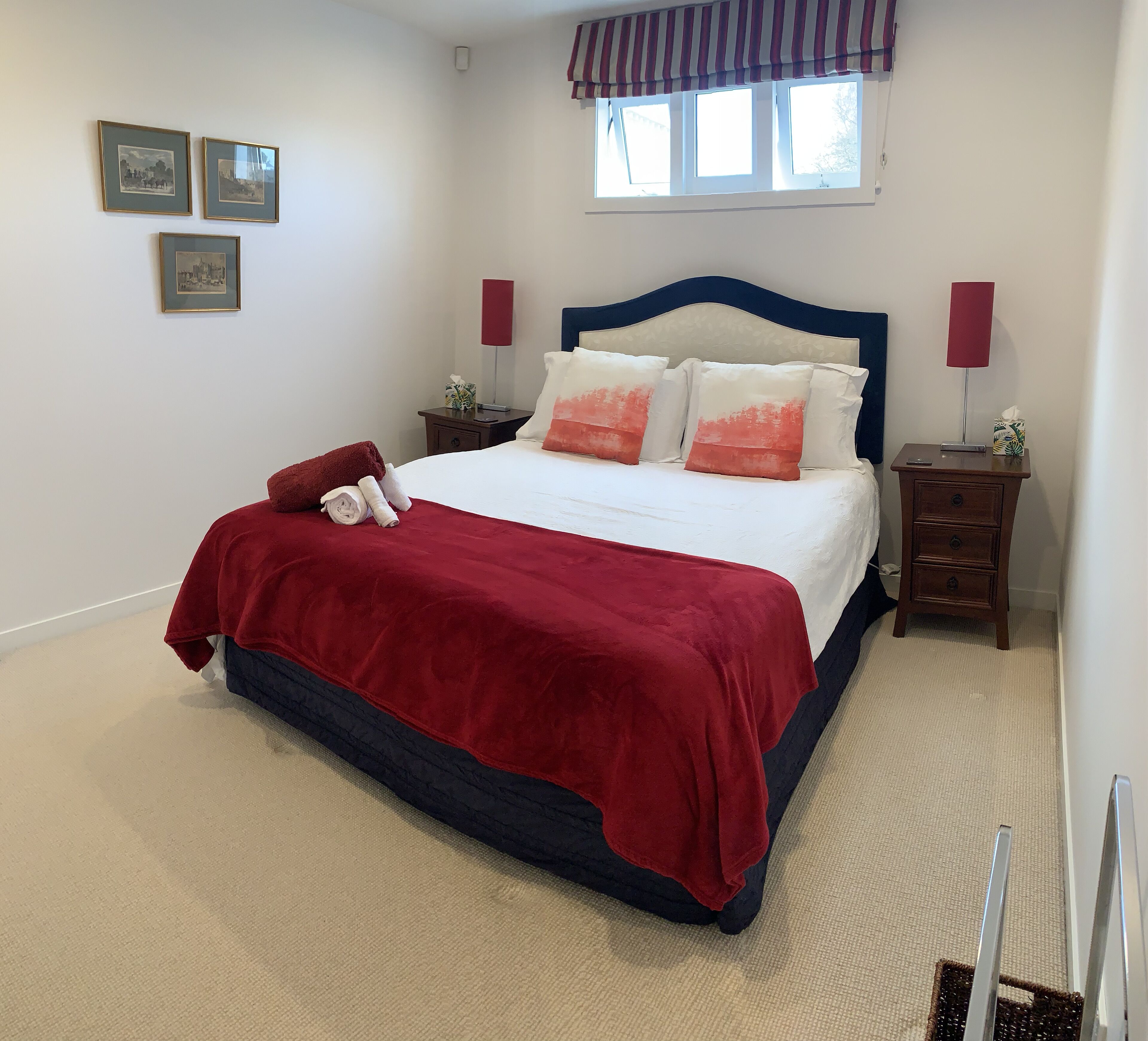 A neatly made bed with a red and blue bedspread, two bedside tables with lamps, and a window with a striped valance.