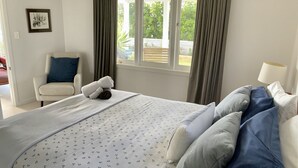 A neatly made bed with a floral-patterned bedspread, a chair with a blue pillow, and a window with curtains.