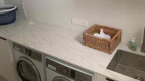 A compact kitchen with a washing machine, a drying rack, a blue laundry basket, and a wicker basket with a bottle of soap.