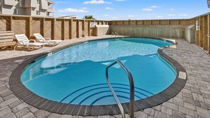 A swimming pool with a wooden deck and lounge chairs, surrounded by a wooden fence and a building with balconies.