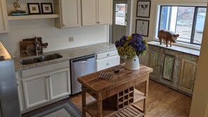 A kitchen with white cabinets, a dishwasher, a wooden table with a vase of flowers, and a pig figurine on the window sill. - Garden Creek Retreat - Remodeled Home with Luxury Furnishings (Challis)