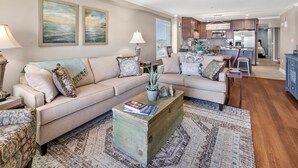 A spacious living room with a large beige sectional sofa, a coffee table with books, and a rug. There is a kitchen in the background with wooden cabinets and a refrigerator. - Upgraded oceanfront condo with pool, kid's pool and private balcony for plenty of sunrises & sunsets (North Myrtle Beach)