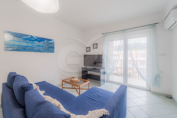 A living room with a blue sofa, a small table, a TV, and a large window with curtains. - Casa Malu apartment 100 meters from the beach (Campo nell'Elba)