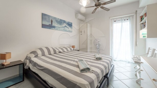 A bedroom with a bed, a ceiling fan, a window with curtains, a bedside table, and a wall-mounted air conditioning unit.