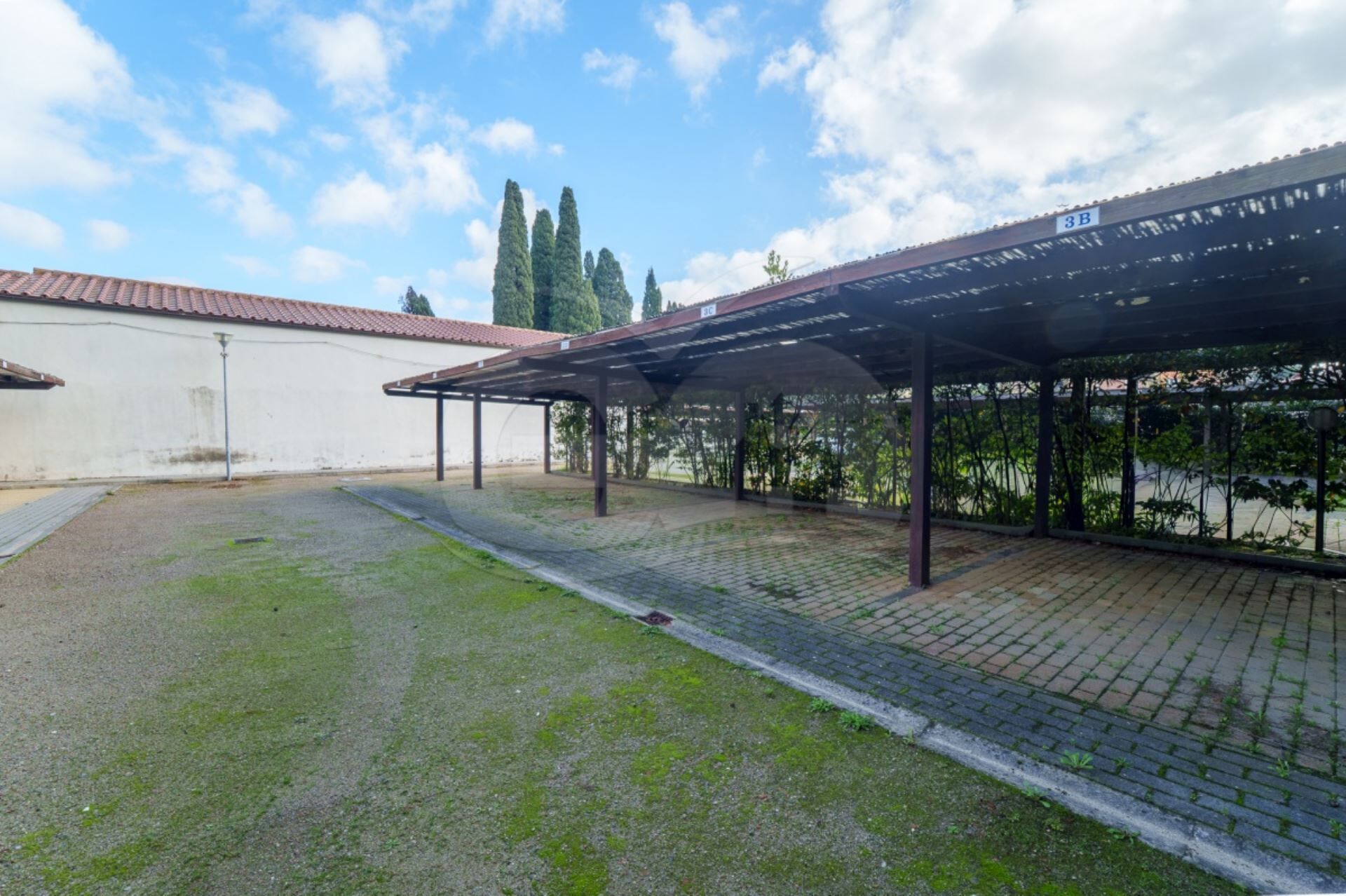 Un espace extérieur abrité doté d’un chemin pavé, un bâtiment blanc avec un toit recouvert de tuiles rouges, et une enseigne indiquant le numéro 3B.