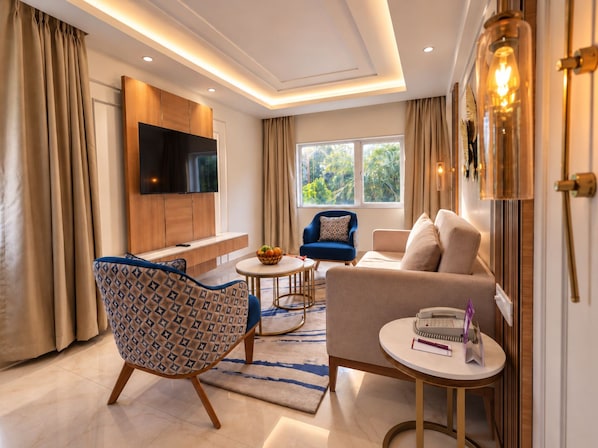 Un salón moderno con un televisor de pantalla plana, un sofá, un sillón azul, una pequeña mesa redonda con un cuenco de fruta y un sillón estampado.