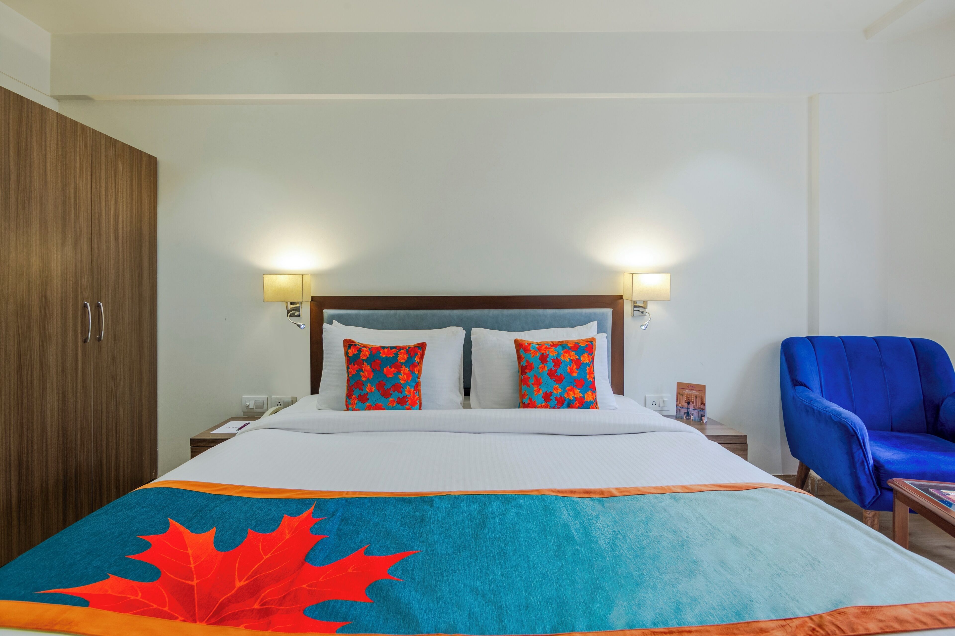 Camera d'albergo con un letto, due cuscini, un comodino, una lampada, una sedia blu e un armadio in legno.