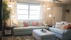 A living room with a light blue sofa, a matching ottoman, a patterned rug, and a large window with curtains. - Orange Tree Cottage- Entire Cottage, Cute and Charming! (Pensacola)