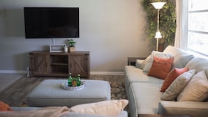 A living room with a flat-screen TV, wooden cabinet, ottoman, and a sofa with cushions. - Orange Tree Cottage- Entire Cottage, Cute and Charming! (Pensacola)