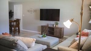 A living room with a sofa, ottoman, and a flat-screen TV on a stand. There is a lamp, a small table, and a cabinet with a tray on top. - Orange Tree Cottage- Entire Cottage, Cute and Charming! (Pensacola)