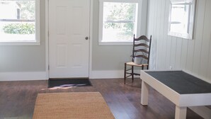 A room with a white door, two windows, a wooden chair, a bench, and a woven rug. - Orange Tree Cottage- Entire Cottage, Cute and Charming! (Pensacola)