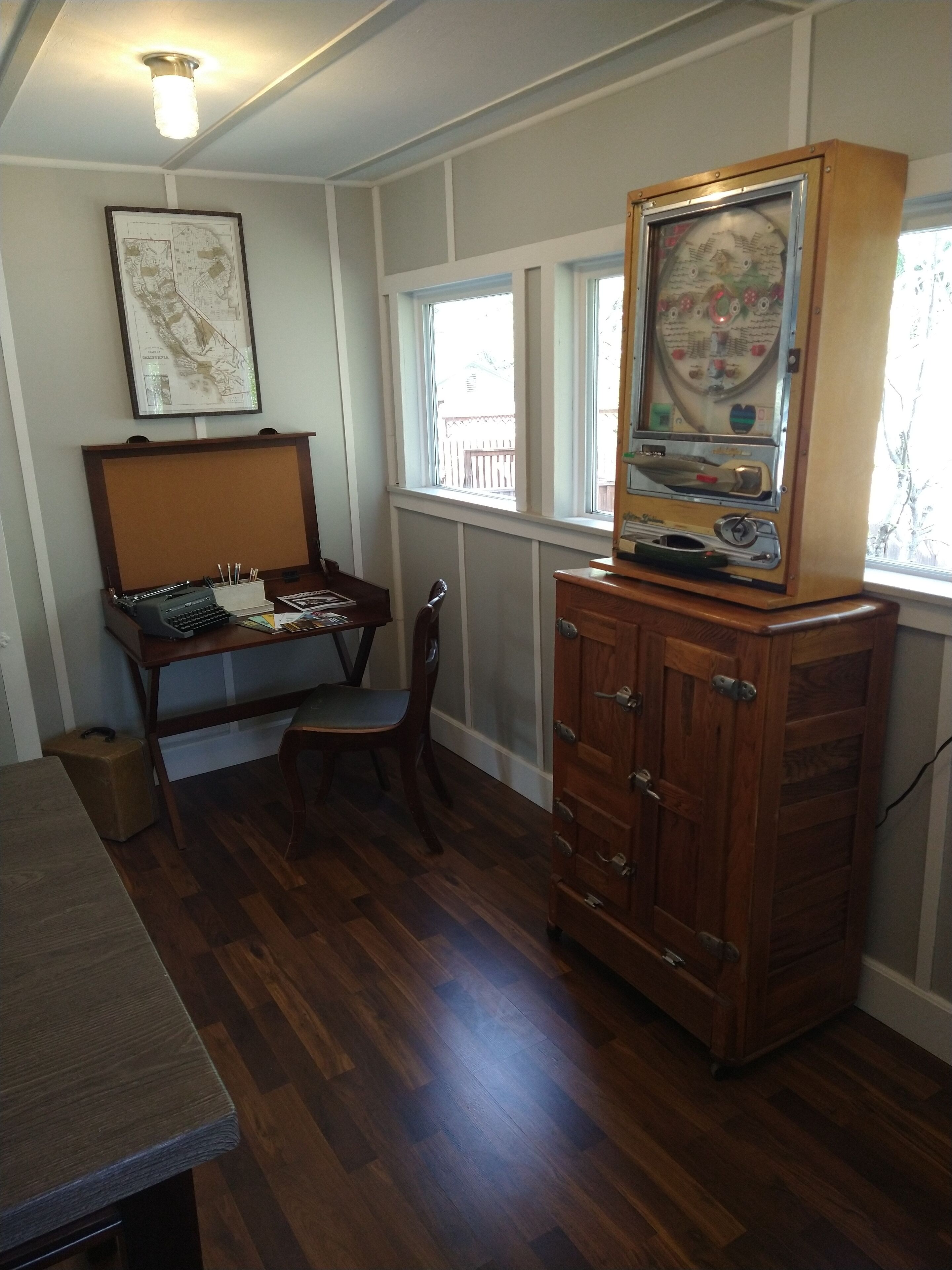 Une pièce avec un bureau en bois, une chaise, un vieux juke-box, une carte accrochée au mur et une fenêtre donnant sur une terrasse.