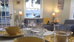 Un coin repas avec des assiettes jaunes et des verres transparents sur une table, un canapé et une fenêtre donnant sur une maison.