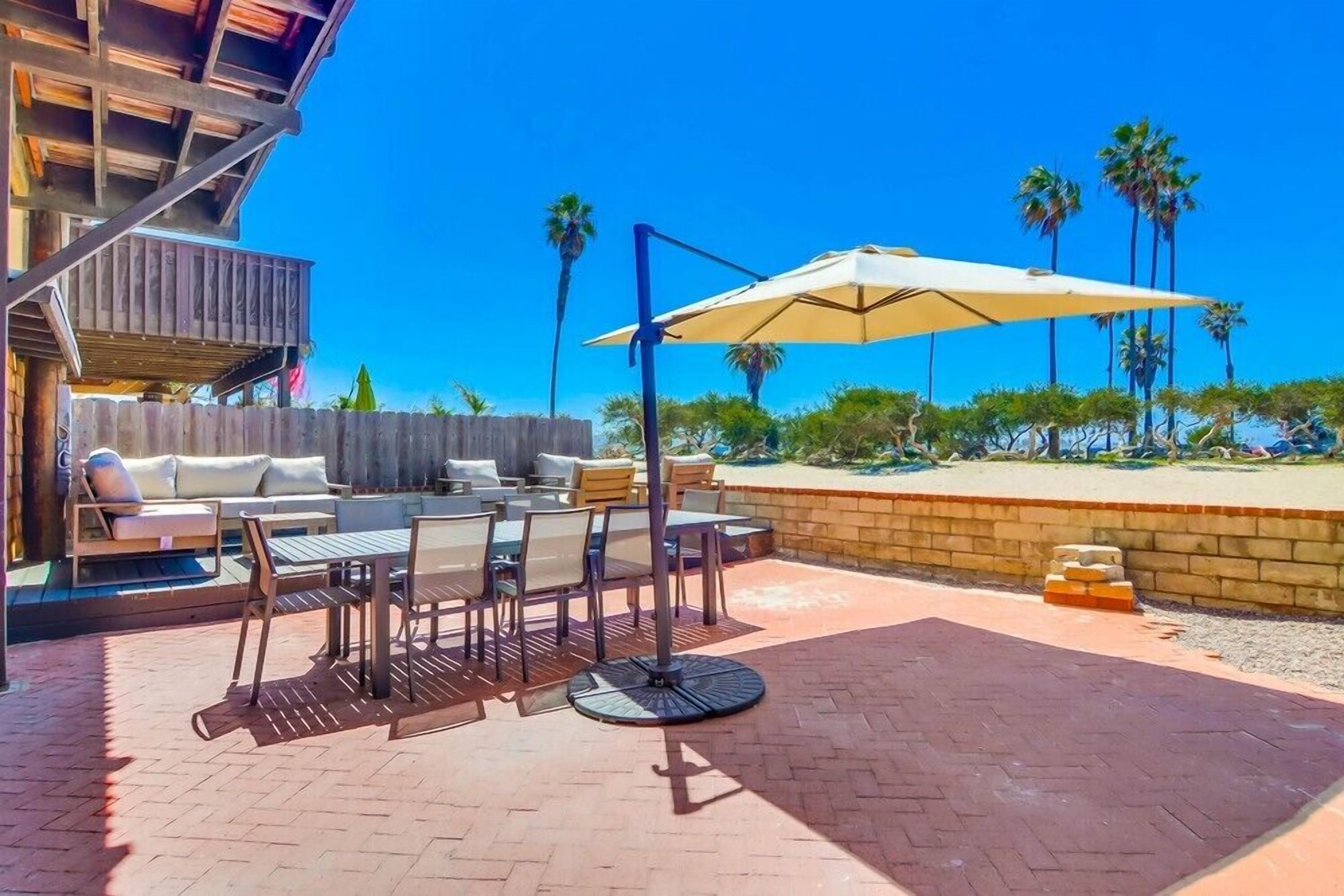 An outdoor patio with a dining table, chairs, and a large umbrella. There is a wooden deck and a brick wall with palm trees in the background.
