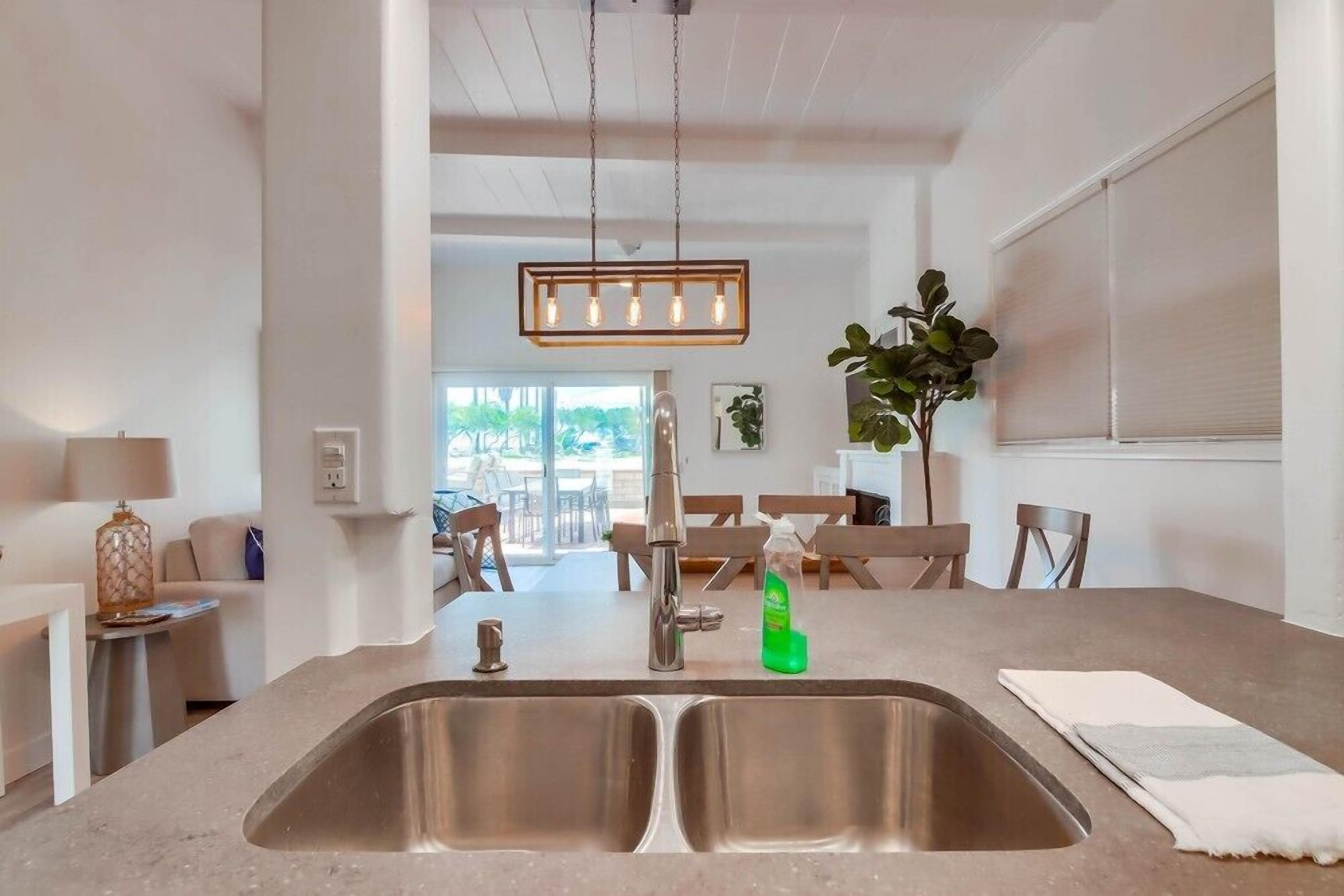 A modern kitchen with a stainless steel sink, a hanging pendant light, and a dining area in the background.