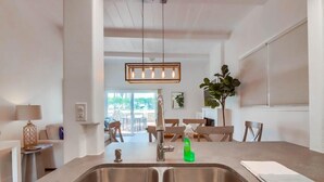 A modern kitchen with a stainless steel sink, a hanging pendant light, and a dining area in the background. - Ocean Front! Exclusive Beachfront Patio + A/C (San Diego)