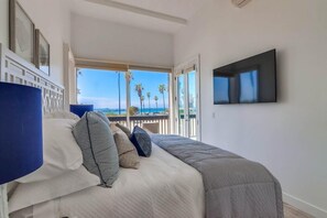 Une chambre à coucher avec un lit, des oreillers, une télévision et un balcon offrant une vue sur les palmiers et la mer.