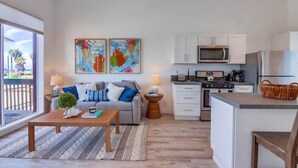 A modern living room with a sofa, coffee table, and artwork on the wall. Adjacent is a kitchen with white cabinets, a microwave, and a refrigerator. - Ocean Front * Panoramic Water View Deck * A/C (San Diego)
