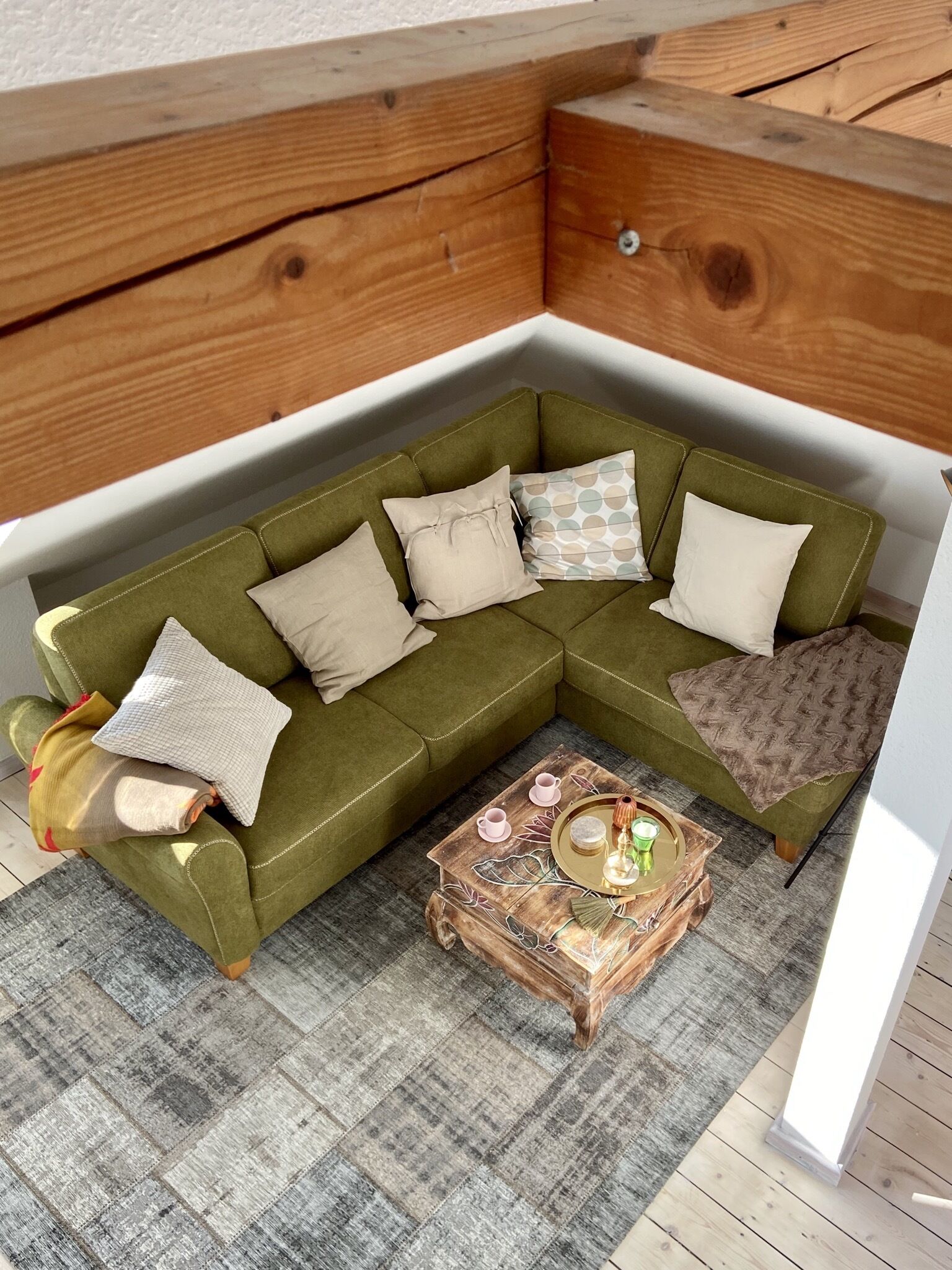 A corner living room with a green sofa, multiple pillows, a wooden coffee table with decorative items, and a patterned rug.