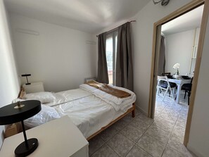 A bedroom with a bed, bedside table, lamp, and a view of a dining area through an open door.