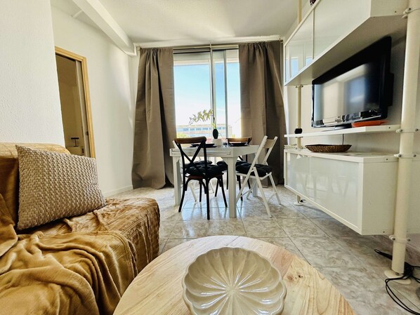 A compact living space with a sofa, a dining table with chairs, a TV on a stand, and a view through the window.