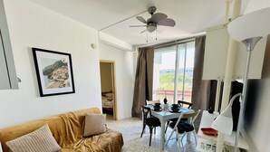 Un salon comprenant un canapé, une petite table, un coin repas avec des chaises, un ventilateur de plafond et une fenêtre avec des rideaux.