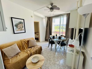 A living room with a sofa, a small table, a dining area with chairs, a ceiling fan, and a window with curtains.