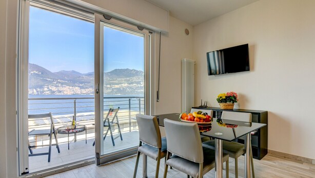 A modern dining area with a glass table and chairs, a flat-screen TV mounted on the wall, and a balcony with a view of the sea and mountains. - Holiday Apartments Brenzone - Apartment SOLE (Brenzone)