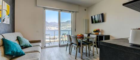 Una sala de estar moderna con sofá, comedor con sillas, mostrador de cocina y un amplio ventanal corredero que da a un balcón con vista a la montaña.