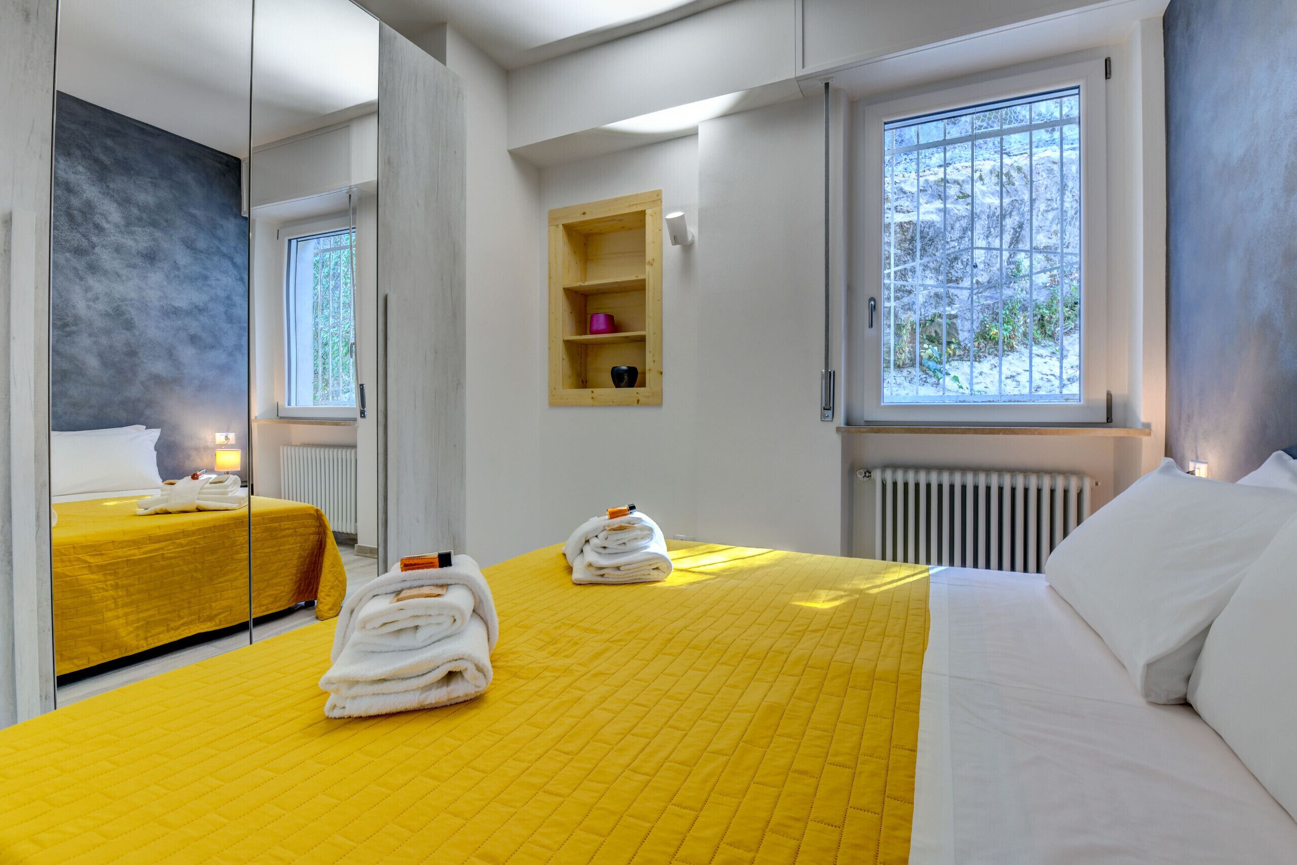 Habitación de hotel con colcha amarilla, un ventanal con vistas a árboles, una pequeña estantería con artículos de aseo y un espejo que refleja la habitación.