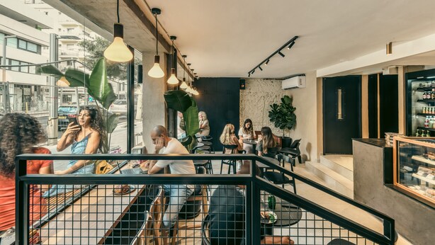 Un café moderne doté d’un escalier menant à un niveau supérieur, de grandes fenêtres, de luminaires suspendus et de différents types de sièges.