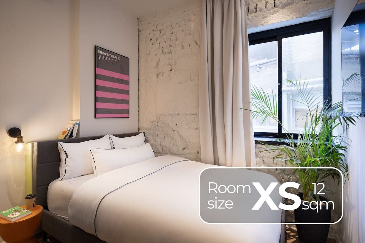 A room with a bed, a nightstand, a potted plant, a window with curtains, and a wall-mounted poster.