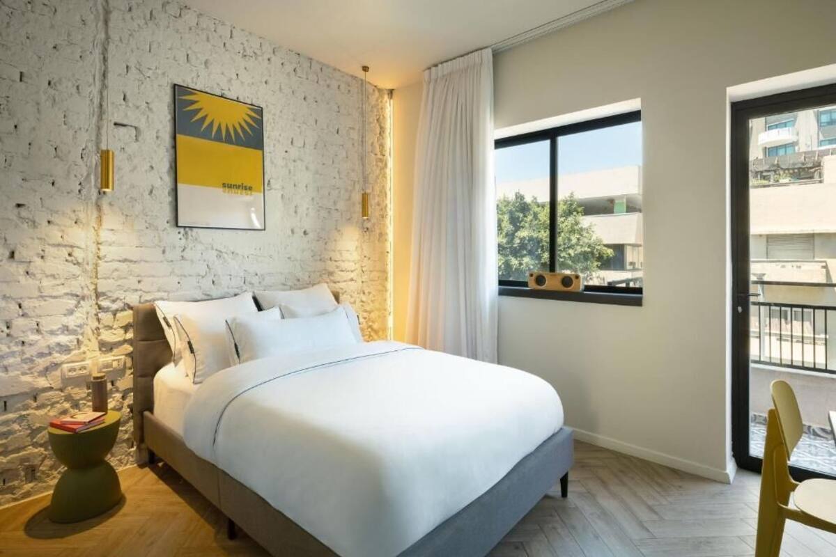 A modern bedroom with a brick wall, a bed with white linens, a window with a view of buildings, and a small table with a chair.