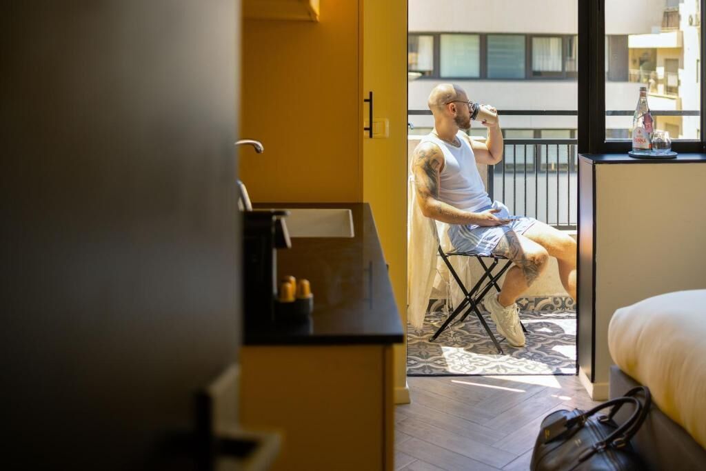 A room with a bed, a chair, a table with a bottle and glasses, and a view of a balcony.