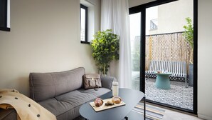 Un salon moderne avec un canapé gris, une petite table ronde sur laquelle se trouvent une tasse de café et des pâtisseries, une plante en pot, et une vue sur un balcon doté d’un sol à motifs et de mobilier d’extérieur.