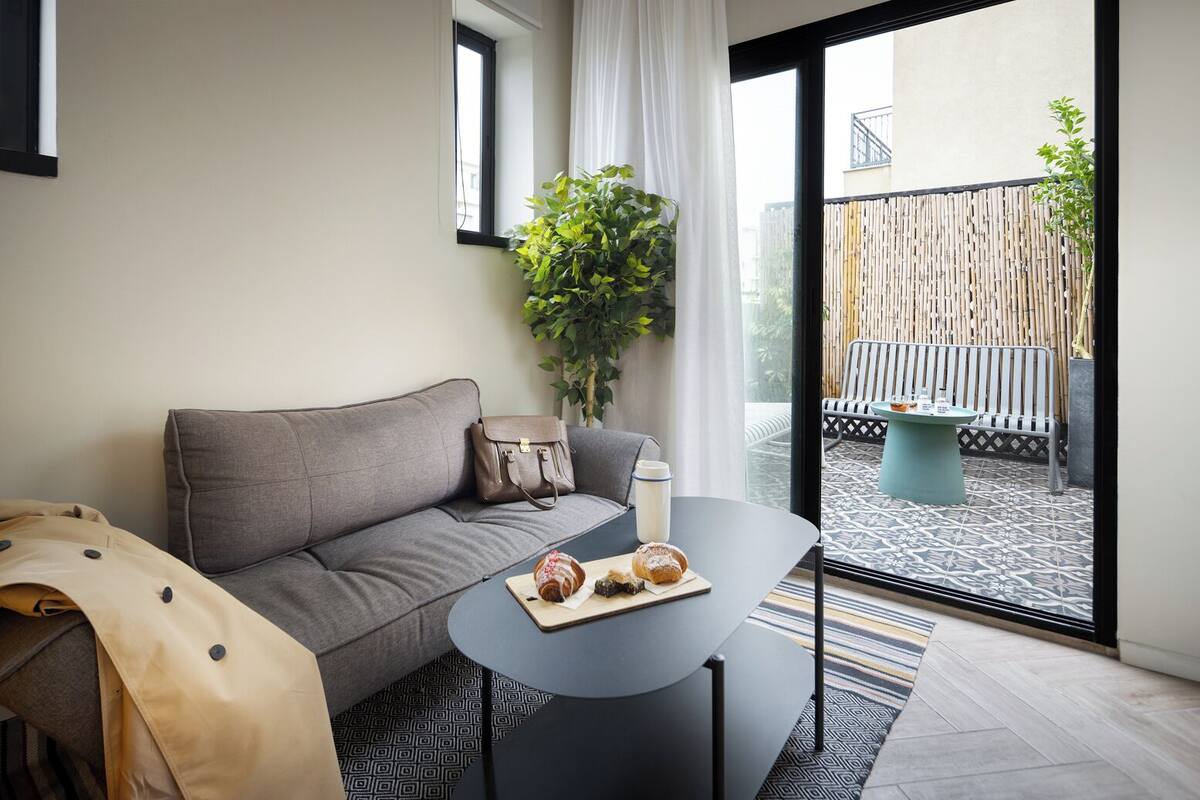 A modern living room with a grey sofa, a small round table with a coffee cup and pastries, a potted plant, and a view of a balcony with a patterned floor and outdoor furniture.