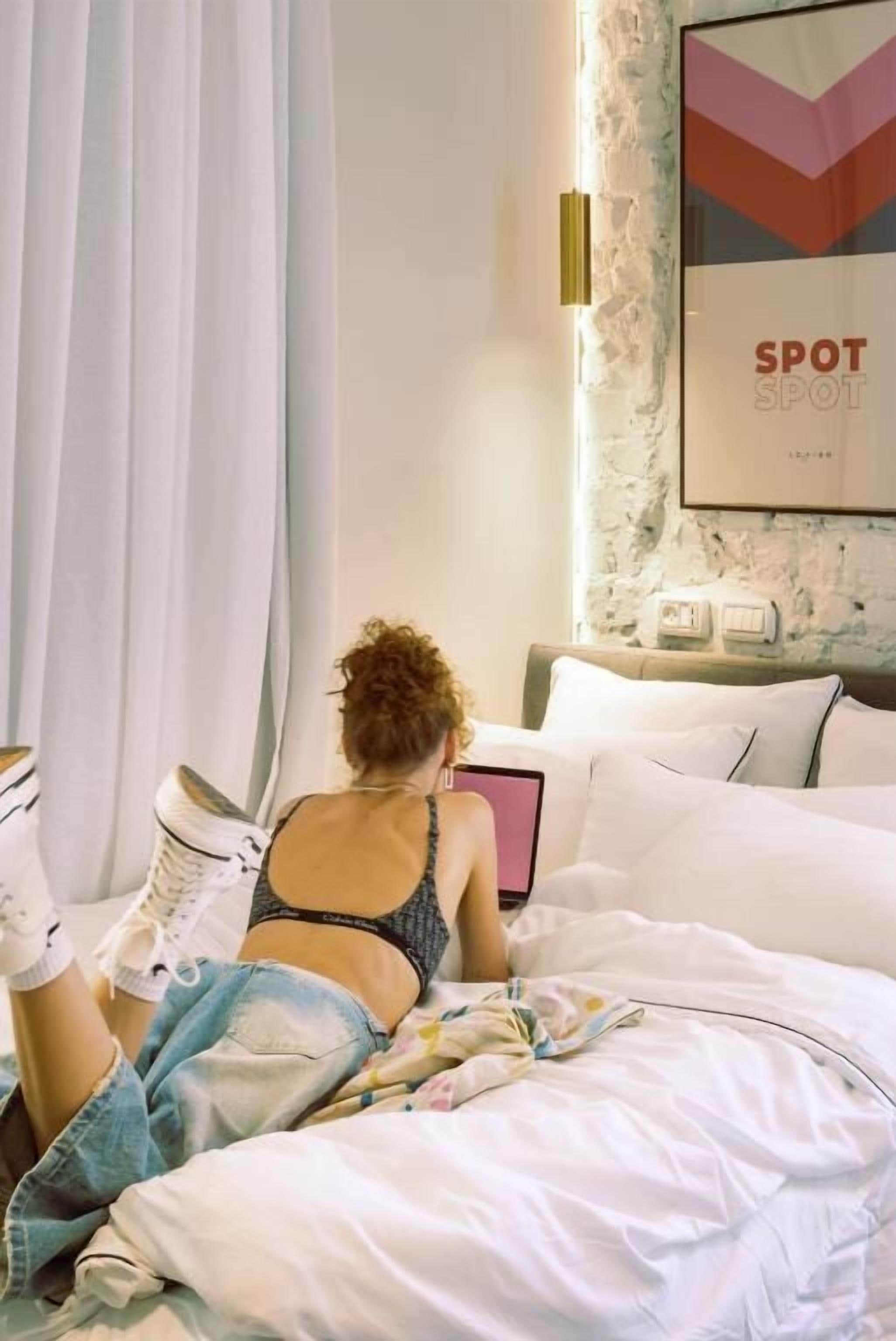 A bedroom with a bed, white curtains, a wall-mounted light, and a framed picture.