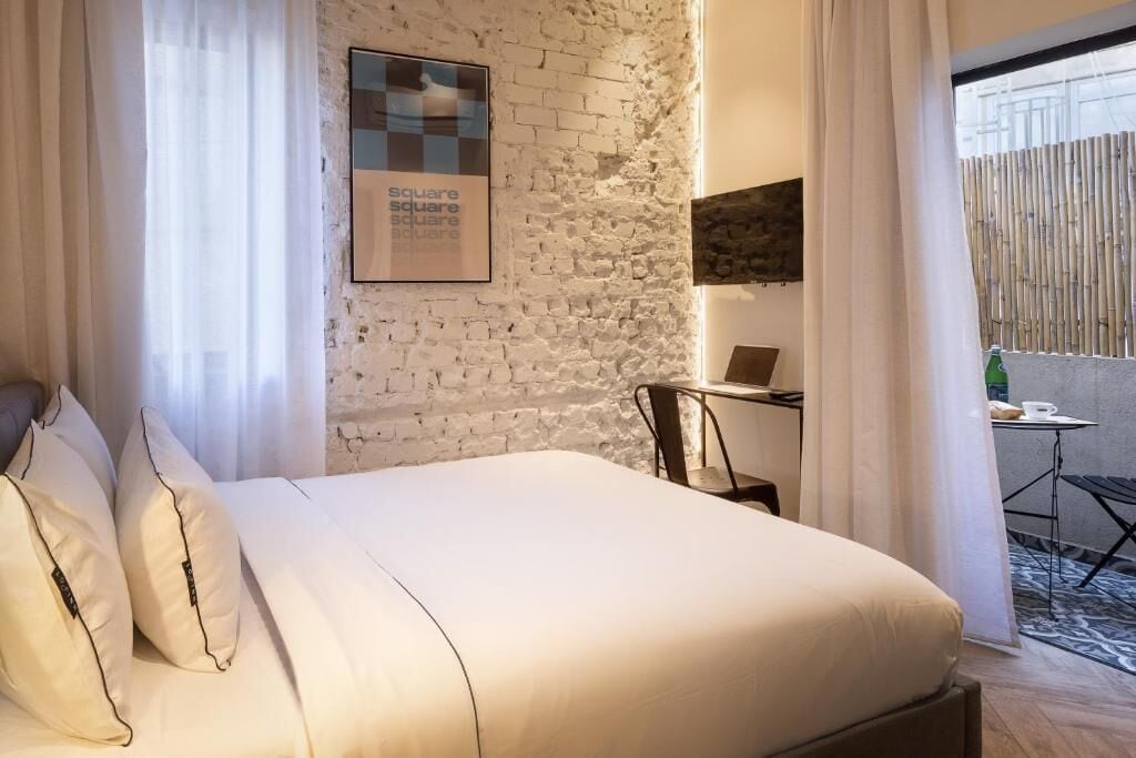 Une chambre avec un mur en briques blanches, un lit avec une literie blanche, un bureau avec un ordinateur portable, et une vue sur une clôture en bambou à l’extérieur.