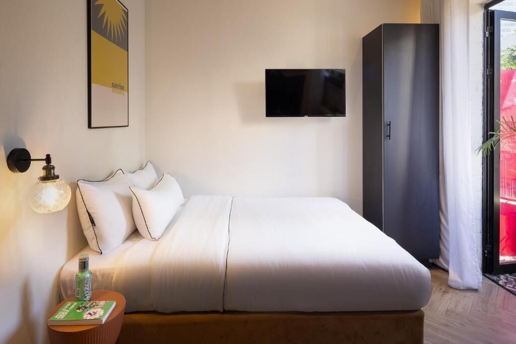 Una cama bien hecha con ropa blanca, un televisor de pantalla plana en la pared, un armario y una mesita con una botella y una revista.