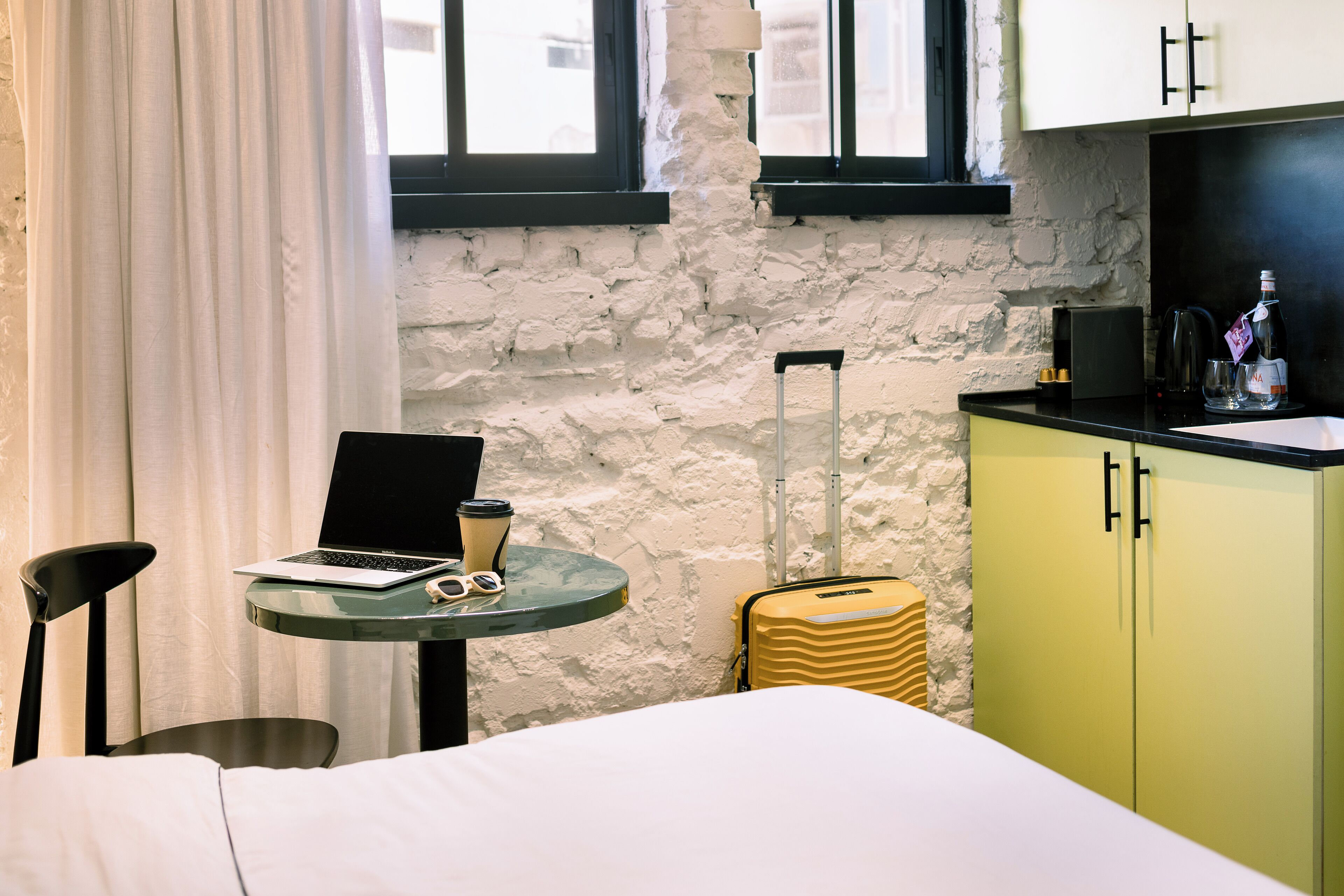 Una habitación pequeña con cama, una mesita con una computadora portátil y una taza, una maleta y una kitchenette.