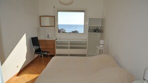 A bedroom with a bed, a chair, a desk, a window with a view of the sea, and a shelf with a lamp.