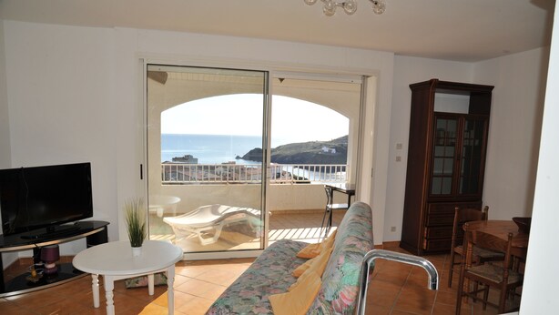 A balcony with a view of the sea, a sofa, a TV, a dining table, and a cabinet.