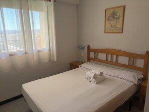 A neatly made bed with white linens and rolled towels, a wooden headboard, a bedside table with a lamp, and a wall-mounted painting of a fruit still life. - Tamaris playa 5ºB (Xeraco Playa)