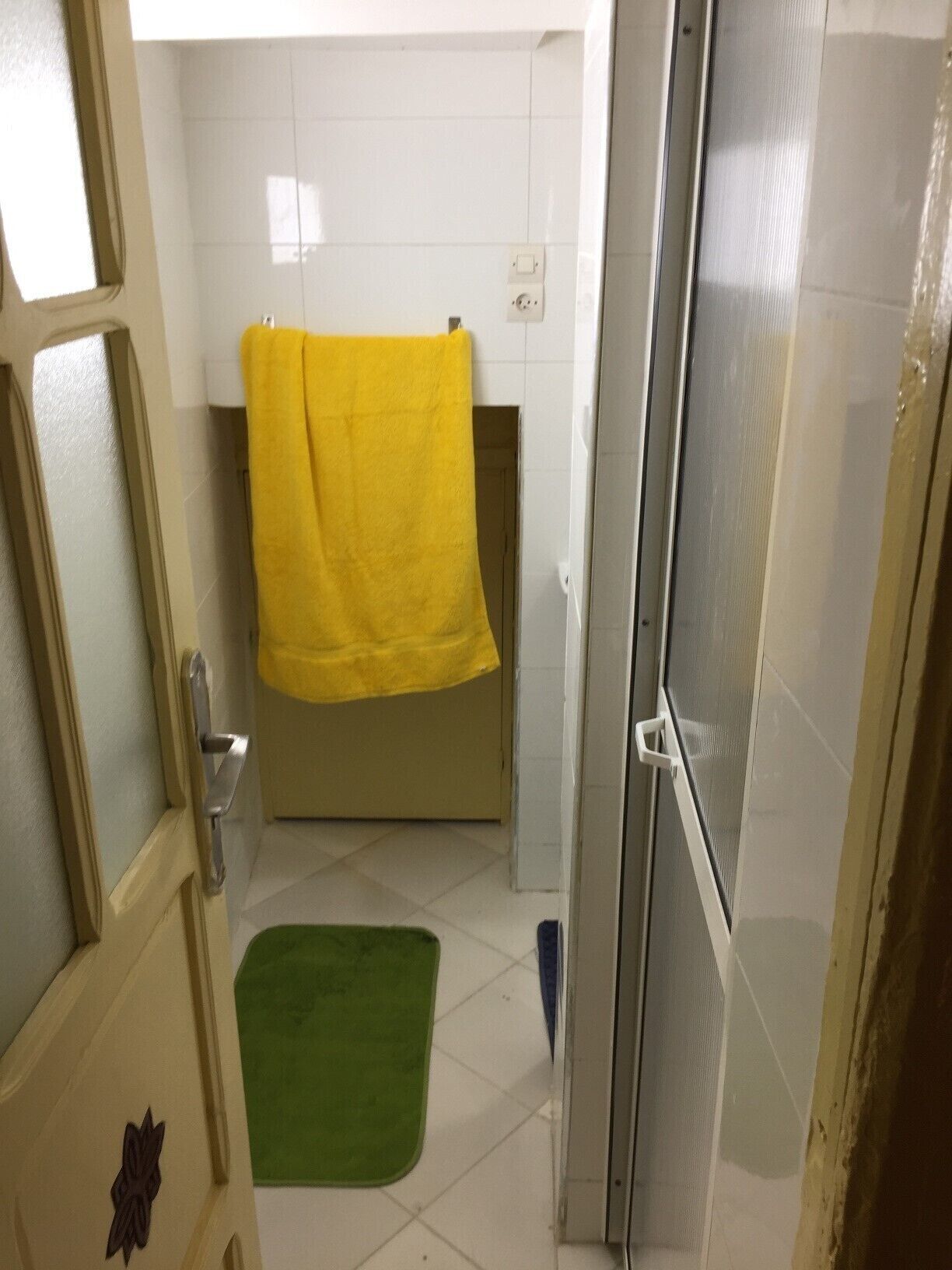 A bathroom with white tiled walls, a yellow towel hanging on a rack, a green mat on the floor, and a shower door.