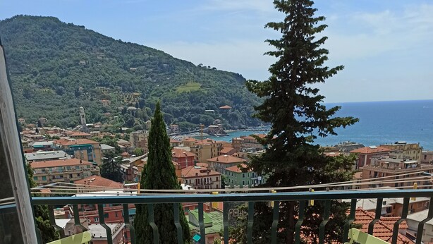 Un balcon avec une table et deux chaises, donnant sur une ville côtière, avec des bâtiments et une colline en arrière-plan.