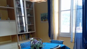 Un coin repas avec une table recouverte d’une nappe bleue, entourée de chaises en bois. On aperçoit, à gauche, un meuble avec des portes en verre, et, à droite, une fenêtre avec des rideaux.