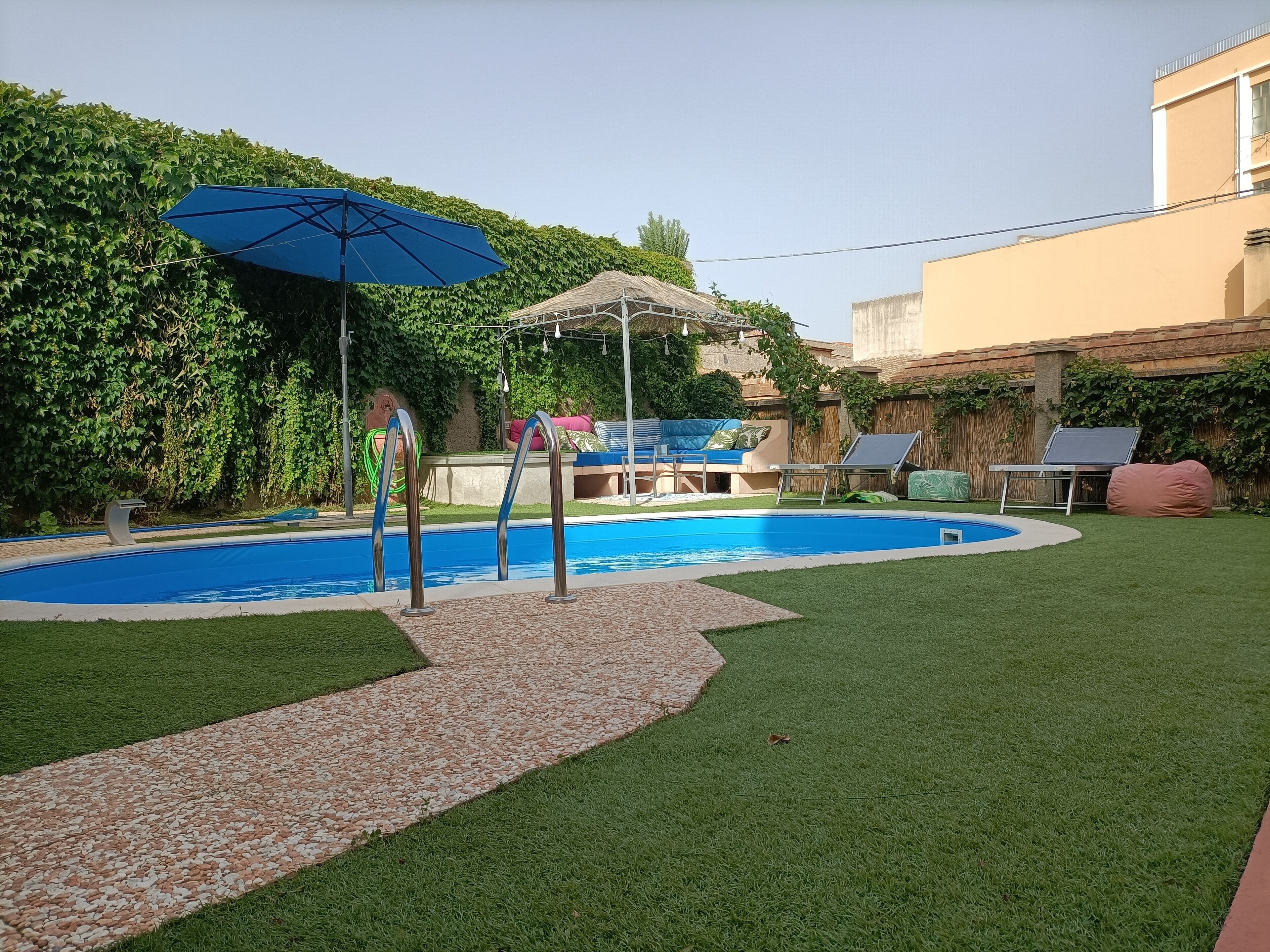 Un jardin avec une piscine, un parasol, un coin salon avec une table et des chaises, et une petite terrasse.